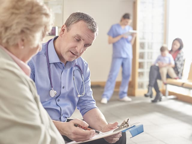 A doctor is talking to a patient in a waiting room with a nurse, child, and woman standing behind them