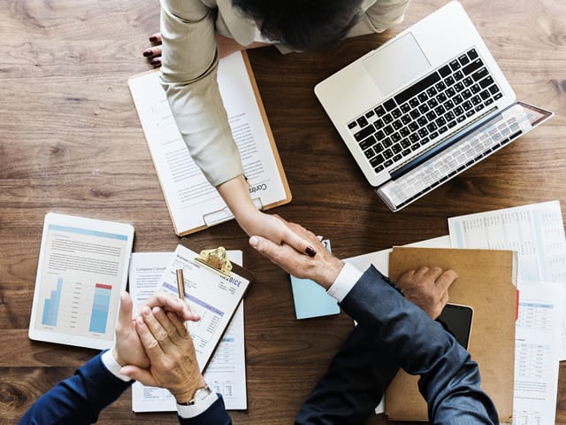 Two people shaking hands while another one claps over a table with a laptop and papers on it