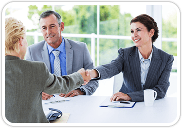 A woman shakes hands with a woman in a suit across the table they are sitting at with another man